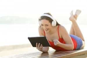 teenage girl wearing headphones watching a video on a tablet on the beach | Online Counseling in Florida | Synergy eTherapy