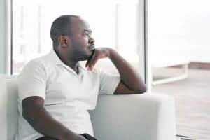 sad african american man sits and looks out the window. He gets online therapy and online grief counseling with Synergy eTherapy